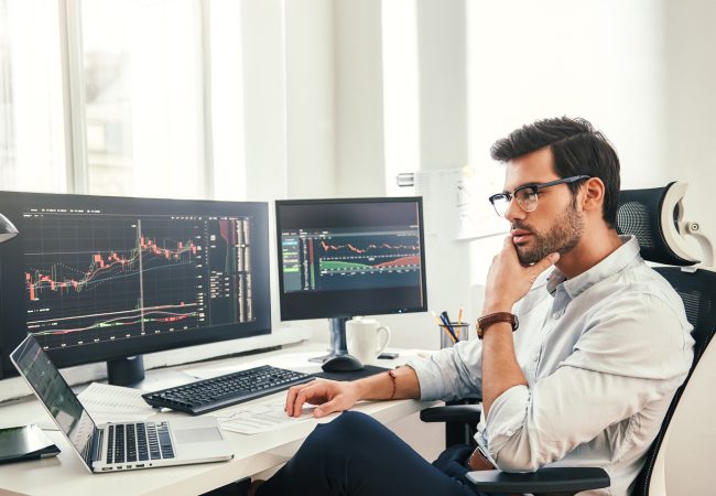 Trading online. Successful and young bearded trader in eyeglasses and formal wear working with laptop while sitting in his modern office in front of computer screens with trading charts. Stock exchange. Trade concept. Investment concept